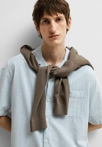 Koszula z guzikami, w niebiesko-białą paskowaną wzór, zestawiona z brązowym swetrem związanym wokół ramion. Gładkie tło.