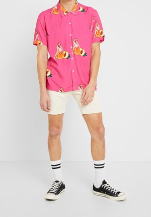 Mann trägt ein hellpinkes kurzärmeliges Hemd mit Knöpfen und einem Muster aus Frauen und Früchten, cremefarbene Shorts, weiße Socken und schwarze Sneaker.