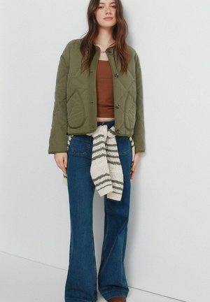 Donna che indossa una giacca trapuntata verde oliva, un top marrone corto, jeans larghi blu e un maglione a righe annodato in vita.