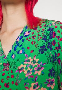 Zielona sukienka w kwiaty z fasonem kopertowym, ozdobiona różowymi, niebieskimi i purpurowymi wzorami kwiatowymi, z teksturowanej tkaniny i dekoltem w kształcie litery V.