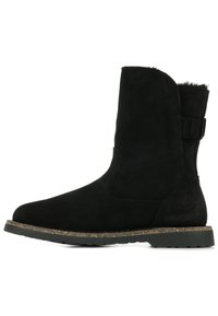 Bota de tobillo de ante negro con punta redondeada, paneles elásticos laterales y suela de goma texturizada. Cuenta con una correa con hebilla en la parte trasera.