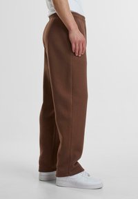 Brune sweatpants med en afslappet pasform, elastisk talje og sidesømme; båret med hvide sneakers, som har en glat, blød tekstur.