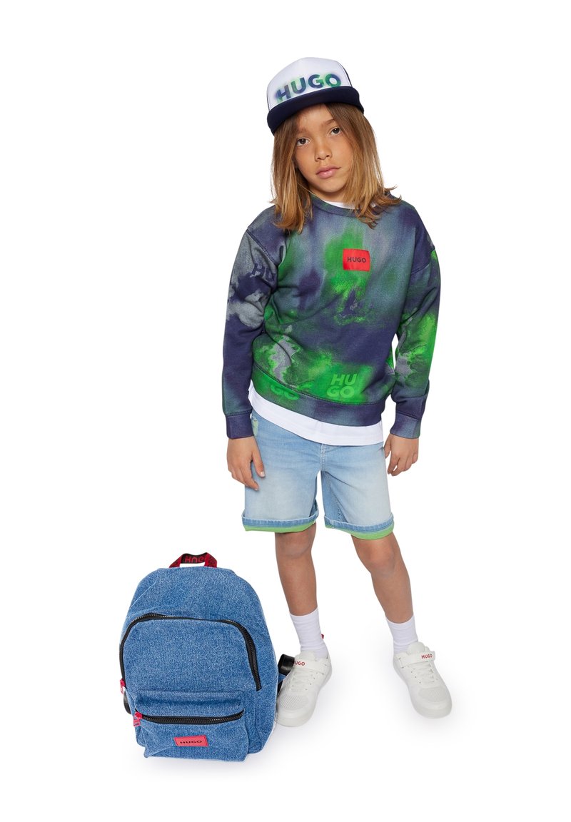 Blue denim backpack with red accents, green and navy patterned sweatshirt with logo, light denim shorts, white sneakers, and a cap.