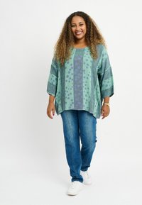 Grön mönstrad topp med blå prickar, oversized passform, trekvartslånga ärmar; i kombination med blå jeans och vita sneakers.