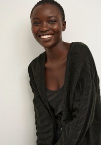 Cárdigan de punto gris oscuro con textura de tejido trenzado, diseño con apertura frontal, usado sobre una camiseta negra. Modelo sonriente frente a un fondo claro.