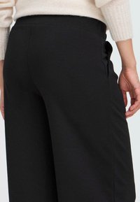 Pantalons noirs à jambes larges en tissu lisse. Dotés d'une taille haute, de poches latérales et de coutures minimales pour un look épuré.