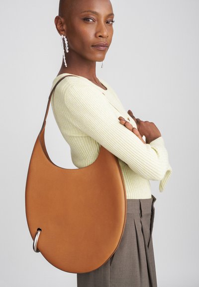 Brūna ādas hobo soma ar izliekta formu, kurai ir sudraba metāla akcenta elements vienā pusē. Gluda tekstūra un minimāls dizains.