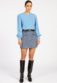 Vrouw in een lichtblauwe lange mouwen sweater, blauwe tweed mini rok met een zwarte riem, en zwarte kniehoge laarzen voor een witte achtergrond.