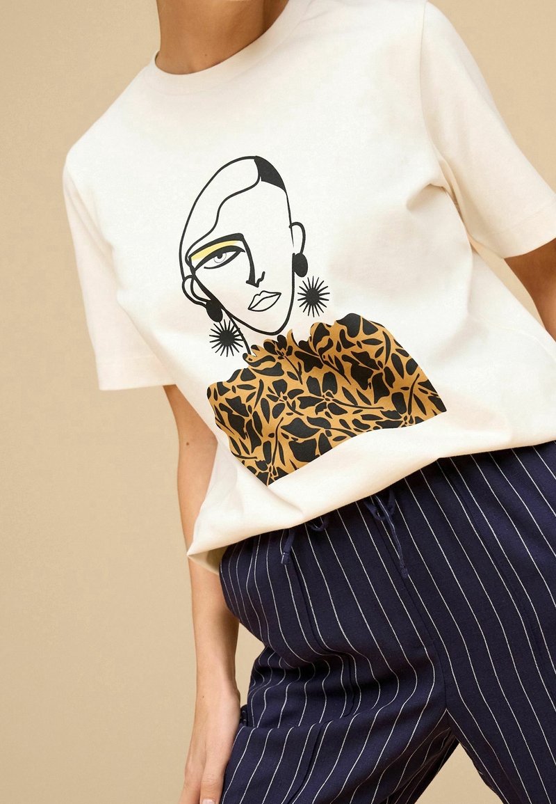 Personne portant un t-shirt blanc avec un visage féminin abstrait et un motif floral, associé à un pantalon bleu marine à fines rayures.
