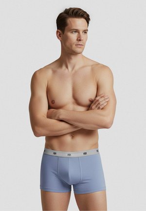 Skjorteløs mand med armene krydset iført lyseblå boxershorts med en grå linning og logo, stående mod en ensfarvet lys baggrund.