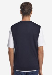 Marinblå stickad väst med rund halsringning, ärmlös design och ribbad fåll. Bärs över en vit t-shirt och visar en slät textur.