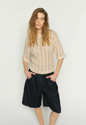 Femeie cu păr lung șaten deschis poartă un pulover tricotat bej și pantaloni scurți negri cu dungi, până la genunchi, pozând cu mâinile în buzunare, în fața unui perete simplu.