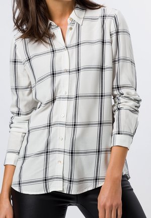 Femme portant une chemise blanche à manches longues avec un motif à carreaux noir et gris et un pantalon noir, debout les mains détendues.
