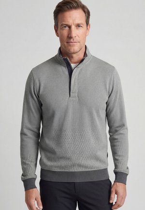 Man draagt een lichtgrijze trui met een kwart ritssluiting, donkergrijze boorden en zoom, gecombineerd met een zwarte broek, staand tegen een effen achtergrond.