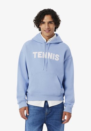 Joven con cabello rizado, que lleva una sudadera con capucha azul claro con el texto "TENNIS", camisa blanca debajo y jeans azules, de pie y sonriendo.