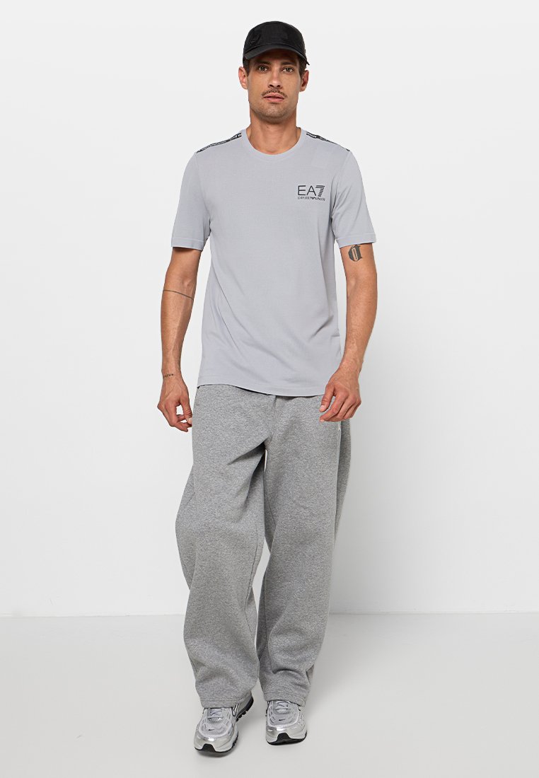 Světle šedé tričko s krátkým rukávem a černým logem, doplněné o volné šedé tepláky. Nošeno s černou kšiltovkou a sportovními botami.