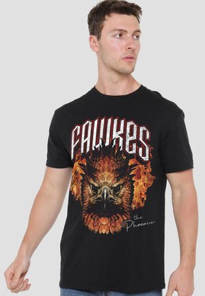 Mann trägt schwarzes T-Shirt mit großer feuriger Phönix-Grafik und Text "FAWKES" und "the Phoenix," und schaut zur Seite vor neutralem Hintergrund.