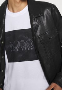 Černá kožená bunda přes bílý tričko s černým obdélníkovým logem s texty "HUGO" a "HUGO BOSS" v tučných písmenech.