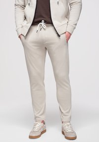 Homme portant un pantalon de jogging beige avec un cordon blanc, un sweat à capuche beige zippé, une chemise marron et des baskets beiges avec des lacets blancs.