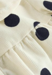 Offwhite krāsas korduro audums ar lieliem melniem polka punktiem. Raksturīga savilkta tekstūra un apakšmala. Mīksta, teksturēta virsma ar redzamu ribojumu.