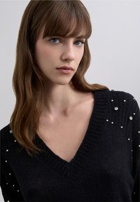 Svart stickad tröja med V-ringning, prydd med pärldetaljer längs axlarna, mjuk textur och ribbade kanter.