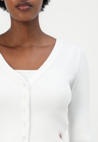 Hvit ribbestrikket cardigan med dyp V-hals, med knapper foran og en liten merkeetikett på nedre høyre side. Mykt, strukturert stoff.