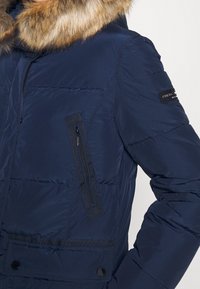 Giacca puffa navy con cappuccio in pelliccia sintetica, tasca sul petto con zip e tasca in vita con bottone a pressione. Texture liscia con design trapuntato.