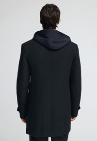 Svart kappa med skräddarsydd design och huva i marinblått. Tyget har en texturerad finish och en strukturerad siluett. Enkla sömnadsdetaljer.