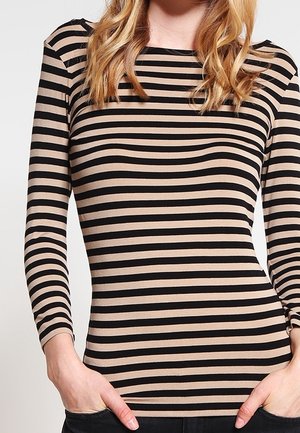 Mujer con una camiseta de manga larga a rayas horizontales en beige y negro, con las manos en los bolsillos de un pantalón negro.