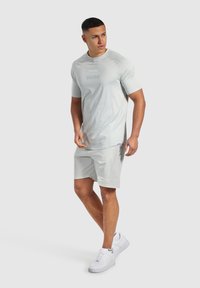 Jasnoszary t-shirt sportowy z krótkim rękawem i nadrukowanymi detalami, w połączeniu z dopasowanymi szortami. Model nosi białe trampki. Gładka faktura.