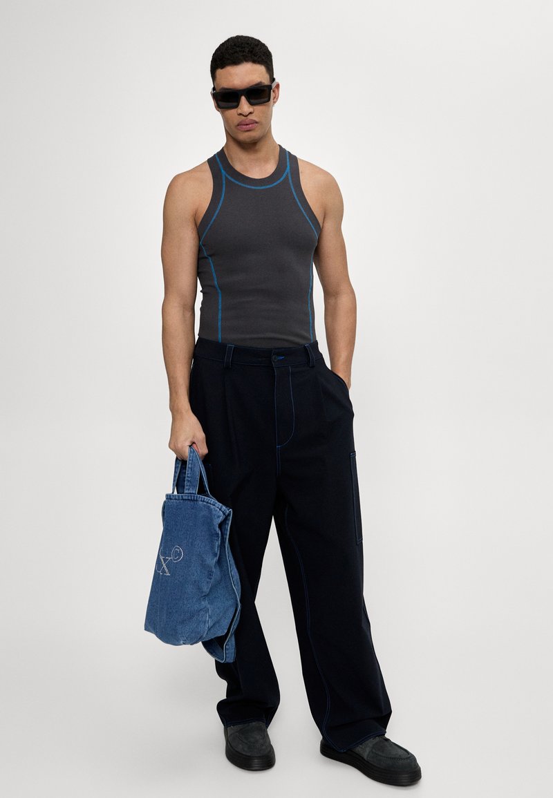 Graues ärmelloses Oberteil mit blauen Nähten, lockere dunkle Hose und eine blaue Jeans-Tasche. Das Modell trägt schwarze Sonnenbrillen und dunkles Schuhwerk.