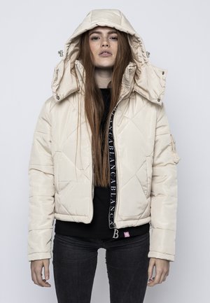 Giacca imbottita crema con cappuccio. Design trapuntato, chiusura frontale con zip e dettagli con logo. Abbinata a una maglietta nera e jeans scuri.