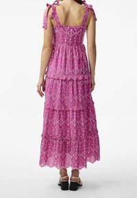 Vestido maxi en encaje rosa brillante con capas en volantes. Presenta bordados decorativos en blanco y lazos ajustables en los hombros. Diseño de espalda cuadrada.