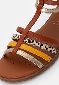 Ruskea tekonahkainen sandaali, jossa on moniväriset remmit: leopardikuvio, metallinhohtoinen kulta, keltainen ja oranssi. Varustettu pehmustetulla pohjallisella, jossa on ilmanvaihtoaukkoja.