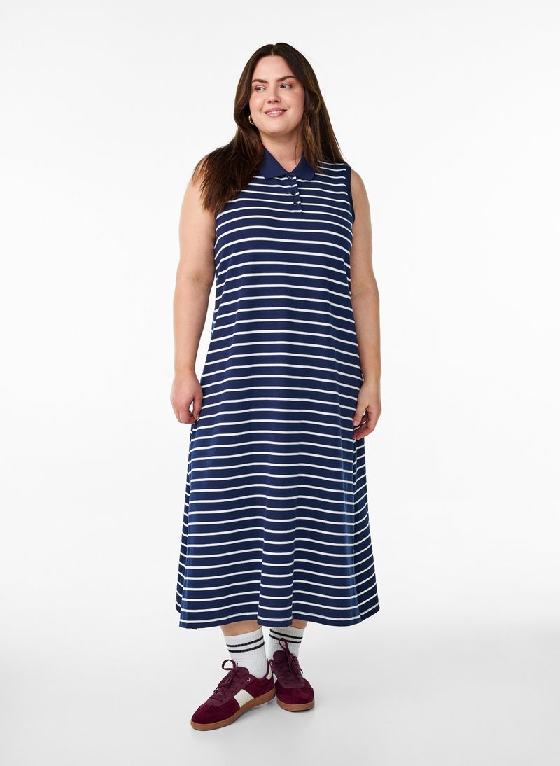 Donna con vestito blu navy senza maniche a righe bianche orizzontali, calzini bianchi e sneakers color granata, in piedi su sfondo bianco.