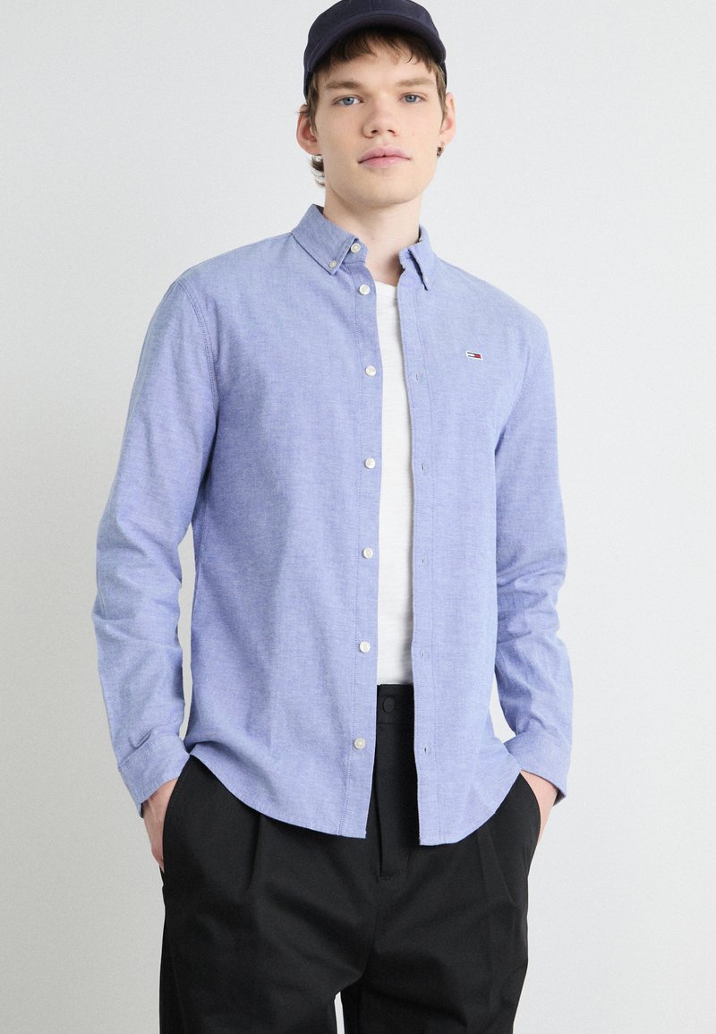 Camisa de botón de color azul claro hecha de tejido entrelazado, con cuello, mangas largas y un pequeño logo en el bolsillo izquierdo del pecho. Pantalones negros.