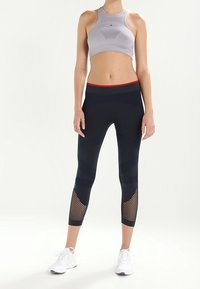 Grå sportbh och svarta capribyxor med röd midja; byxorna har meshpaneler och texturerade ränder. Vita sportskor.