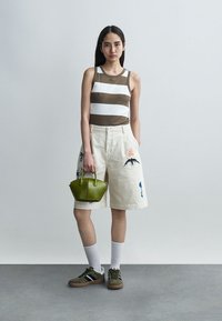 Jeune femme portant un débardeur rayé marron et blanc, un short crème brodé arrivant aux genoux, tenant un sac à main vert, chaussée de baskets et de chaussettes blanches.