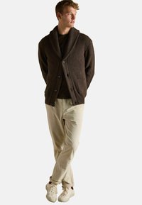 Jauns vīrietis stāv ar rokām aiz muguras, ģērbts tumši brūnā šallē ar apkakli, melnā kreklā, bežās biksēs un baltos sporta apavos.