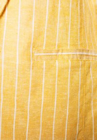 Żółty len z białymi pionowymi paskami i widoczną kieszenią. Materiał ma fakturowane sploty, co zwiększa atrakcyjność wizualną.