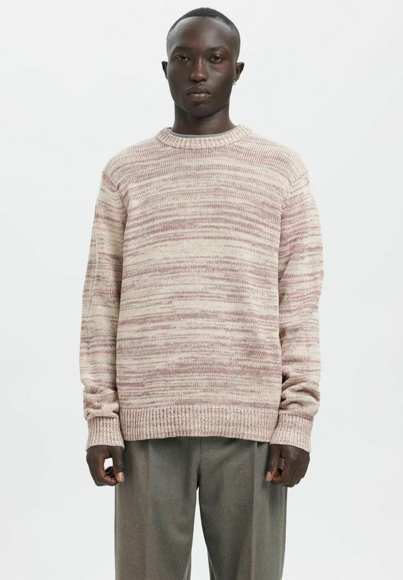 Maglione in morbido beige con righe rosa, caratterizzato da scollo e polsini a coste. Vestibilità comoda con spalle scese. Indossato con pantaloni grigi.