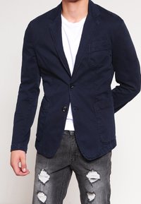 Blazer in cotone blu navy con design a petto singolo, revers a punta, due tasche frontali e una tasca sul petto, abbinato a jeans grigi effetto vissuto.