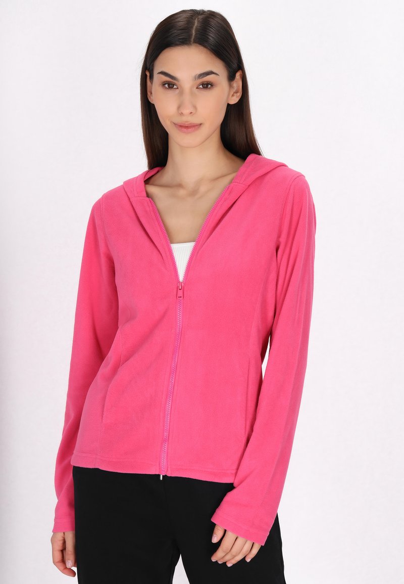 Felpa in pile di un vivace rosa, con chiusura a zip, cappuccio, maniche lunghe e una texture morbida, abbinata a una maglietta bianca e pantaloni neri.