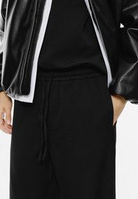 Persoon in zwarte drawstring broek, zwart shirt, gestreept wit overhemd en zwarte leren jas met één hand in de zak.