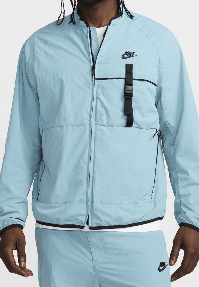 Ljusblå Nike-jacka med dragkedja framtill, svarta accenter och bröstficka. Har en slät textur och en sportig design.