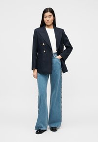 Γυναίκα που φοράει σκούρο μπλε διπλόμπουτο σακάκι, λευκό πουκάμισο, φαρδιά μπλε τζιν με διακοσμητικά στη μία πλευρά και μαύρα μοκασίνια, όρθια με το ένα χέρι στην τσέπη.