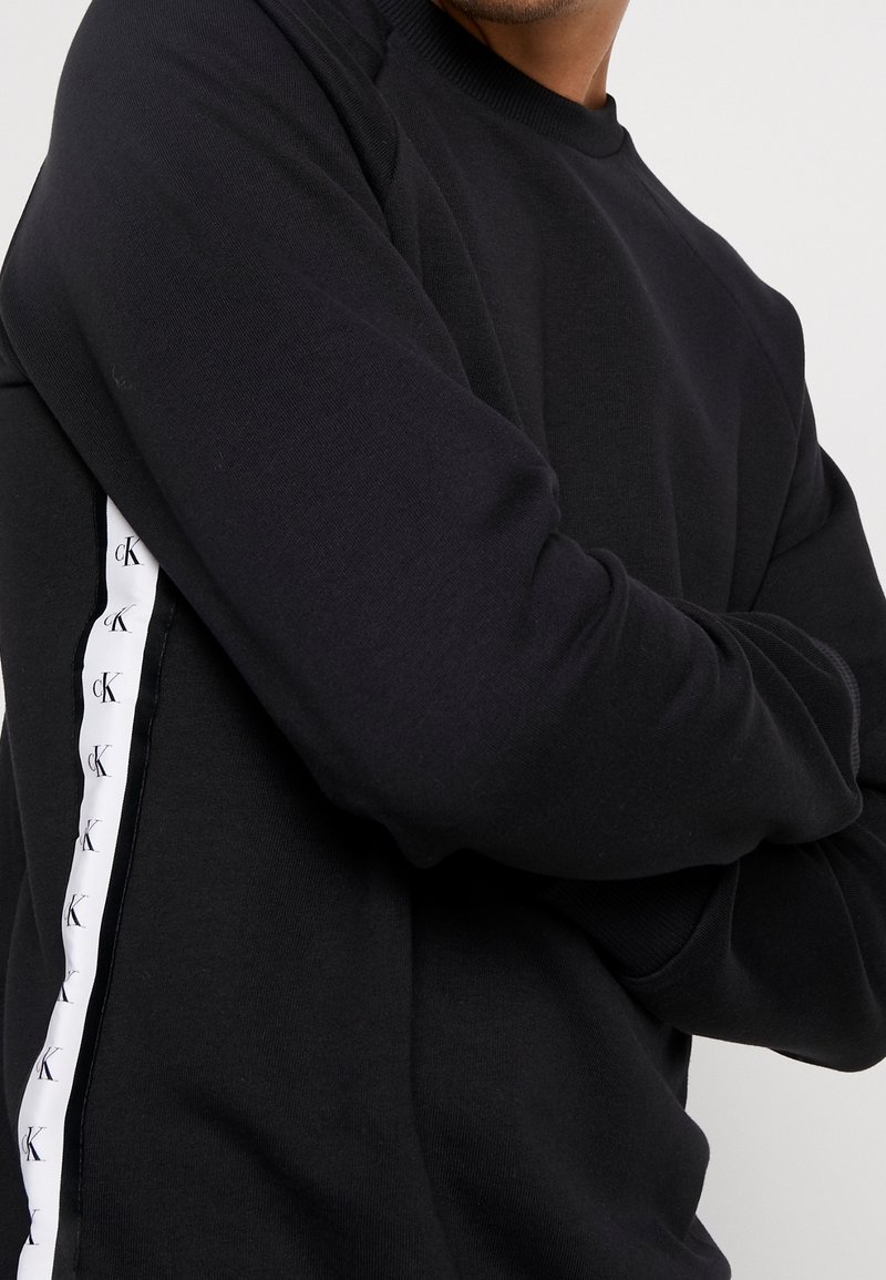 Sudadera negra con cuello redondo, con una franja lateral blanca adornada con un logotipo repetido. El material parece suave y cuenta con puños acanalados.