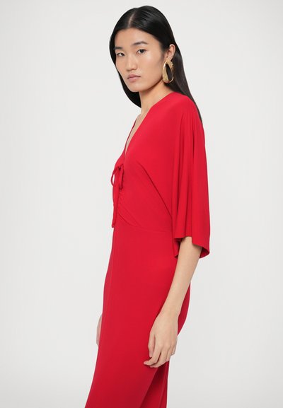 Sarkana plūstoša kleita ar V veida izgriezumu un saišu detaļu vidū. Aprīkota ar īsām piedurknēm un gludu, reljefainu audumu. Zelta riņķveida auskari.