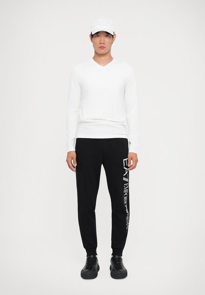 Cămașă albă cu mâneci lungi și decolteu în V, pantaloni de trening negri cu text alb, adidași negri și o șapcă albă, stând în fața unui fundal simplu.