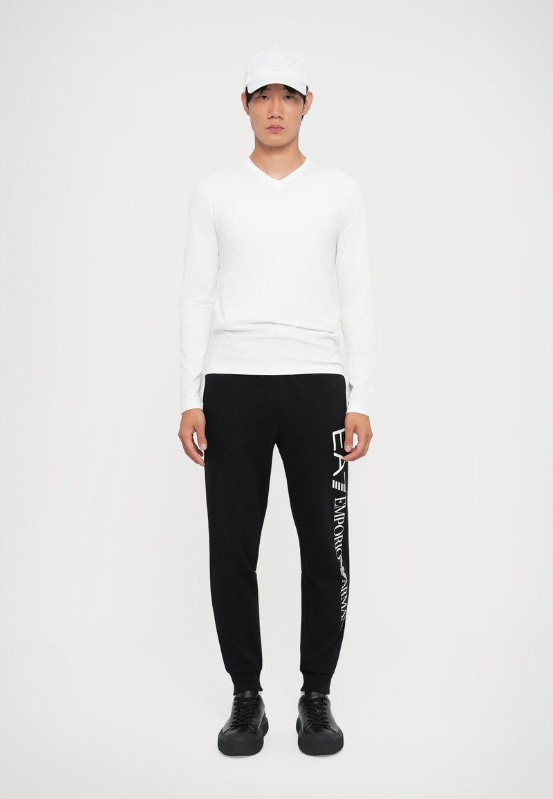 Camisa blanca de manga larga y escote en V, pantalones de chándal negros con texto blanco, zapatillas negras y una gorra blanca, de pie contra un fondo simple.
